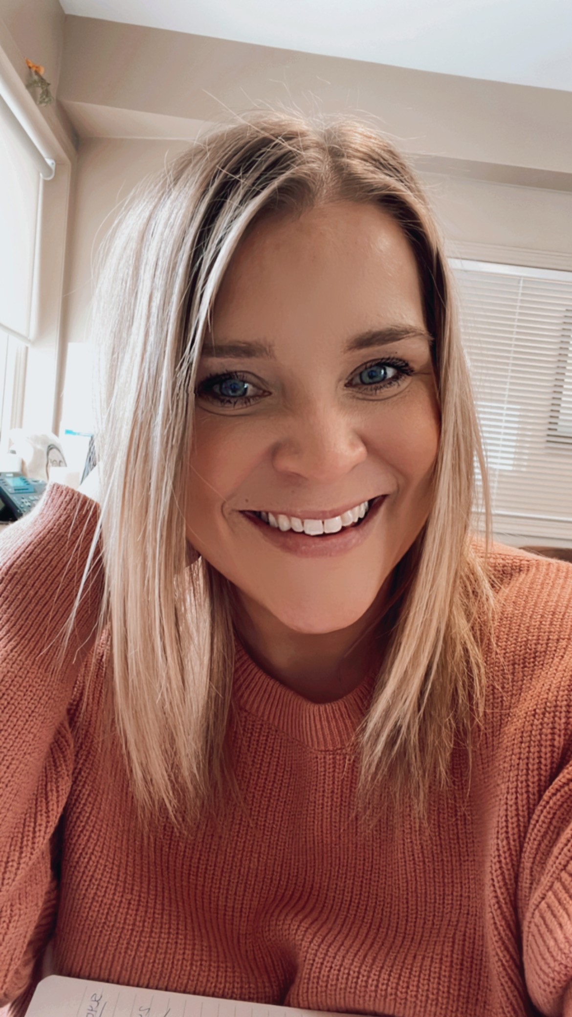 Woman, smiling, straight blonde hair, peach shirt, leaning on right hand, sitting 