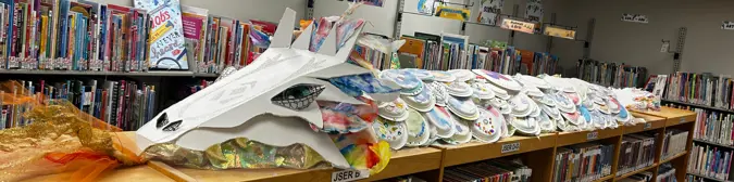 A large multi-coloured dragon constructed of paper plates lies across the top of some book shelves.
