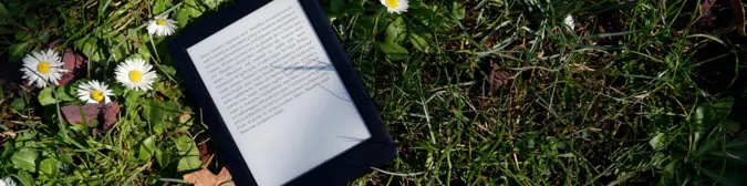 An eReader laid out in the grass with daisies to the left of it.