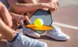 Close up of two people sitting down on ground holding pickleball paddles and ball