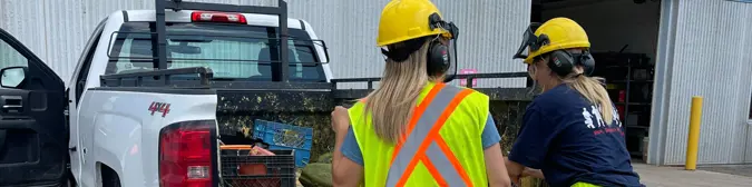 [Two summer students wearing hardhats and removing lawn care tools from a truck.'